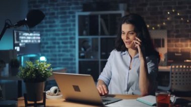 Neşeli kadın cep telefonu konuşuyor ve karanlık ofiste dizüstü bilgisayar ile çalışıyor