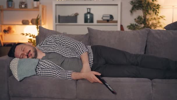 Портрет человека, дремлющего на диване дома ночью с дистанционным управлением в руке — стоковое видео