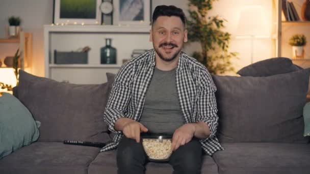 Ragazzo allegro che guarda la TV che ride indicando lo schermo e mangiando popcorn a casa — Video Stock