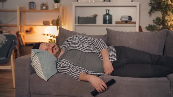 Jeune homme dormant sur le canapé à la maison tenant télécommande pour la télévision — Video