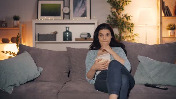 Ung dam tittar på TV och äta popcorn hemma sitter på soffan ensam — Stockvideo