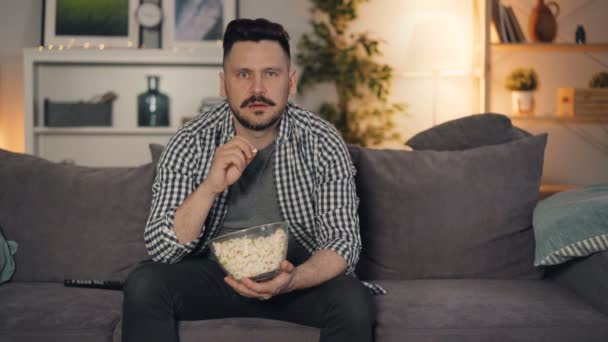 Joven barbudo viendo televisión y comiendo palomitas en casa sentado en el sofá — Vídeos de Stock