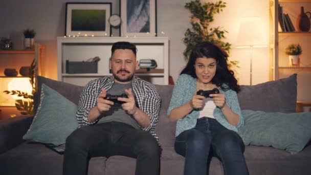 Esposo y esposa jugando videojuego sosteniendo joysticks sentado en el sofá en casa — Vídeos de Stock