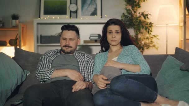 Муж и жена смотрят драму по телевизору с грустными лицами, сидящими на диване в доме — стоковое видео