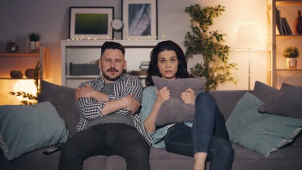 Esposo y esposa viendo películas de miedo en la televisión mirando a la cámara con caras asustadas — Vídeos de Stock