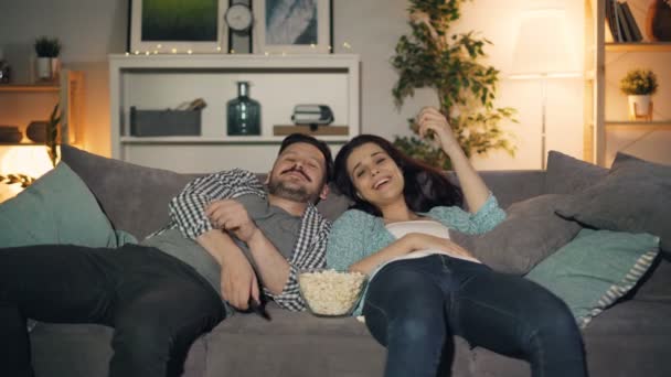 Χαρούμενοι άνθρωποι που κοιτάζουν την τηλεόραση γελώντας τρώγοντας ποπ κορν ξαπλωμένοι στον καναπέ στο σπίτι — Αρχείο Βίντεο
