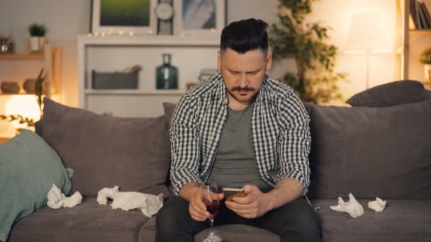 Депрессивный парень с помощью смартфона держит стекло с алкоголем сидя дома в одиночестве — стоковое видео