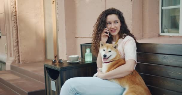 Девушка разговаривает по мобильному телефону и обнимает шибу-ину собаку на улице в кафе — стоковое видео