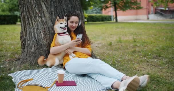 Modern lány zenét hallgat fejhallgató segítségével okostelefon parkban kutyával — Stock videók