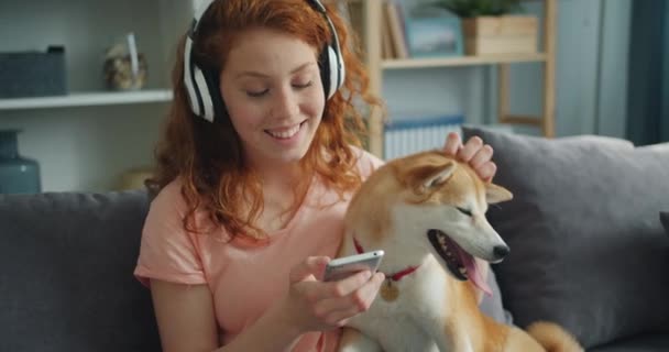 Beautiful woman in headphones listening to music and petting dog on couch — Stock Video