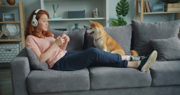 Chica usando teléfono inteligente en auriculares sentados en el sofá en casa con perro mascota — Vídeos de Stock