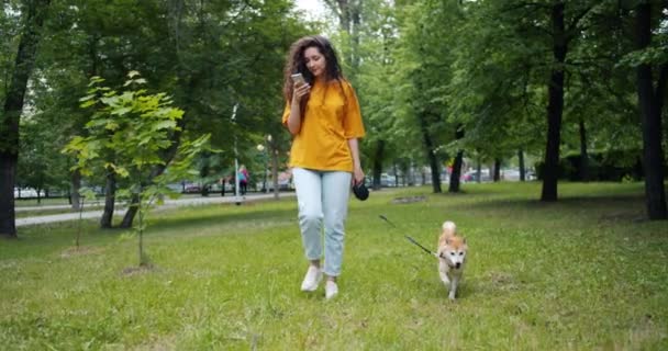 Mooi meisje wandelen mooie Shiba Inu hond in het park met behulp van smartphone buitenshuis — Stockvideo
