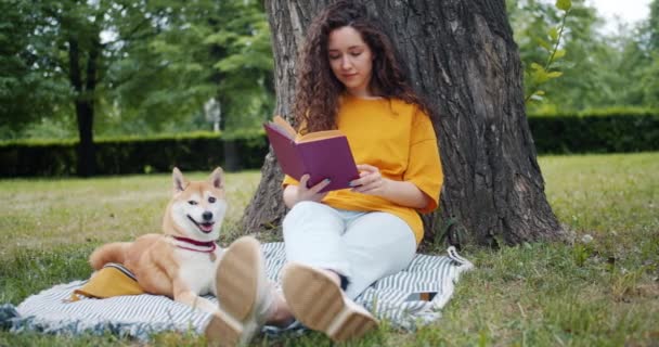 Ładne kręcone włosy uczennice czytanie książki siedzącej w parku na trawniku z psem — Wideo stockowe