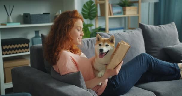 Pěkná studentka četba v bytě usmívající se a peče rozkošný pes — Stock video