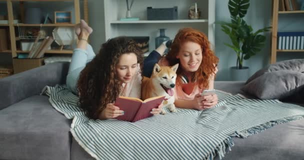 Девушки читают книгу, используя смартфон, лежащий на кровати с собакой в квартире — стоковое видео