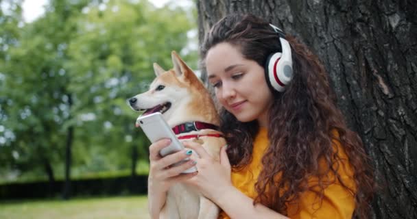 Lachende student genieten van muziek in een koptelefoon met behulp van smartphone in Park met hond — Stockvideo