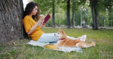 Rahat kız kahve içme ve parkta kitap okuma köpek ile çim üzerinde oturan