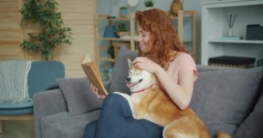 Güzel genç bayan kitap okuma ve evde kanepede sevimli köpek okşayarak