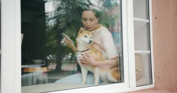Леди слушает музыку с наушниками и смартфоном целует собаку в кафе — стоковое видео
