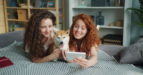 Блудниці беруть селфі з милим щеням, що лежить на дивані вдома, використовуючи смартфон — стокове відео