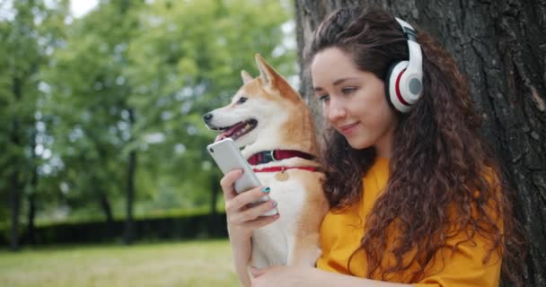 Senhora feliz em fones de ouvido usando smartphone, em seguida, abraçando cão de estimação no parque — Vídeo de Stock