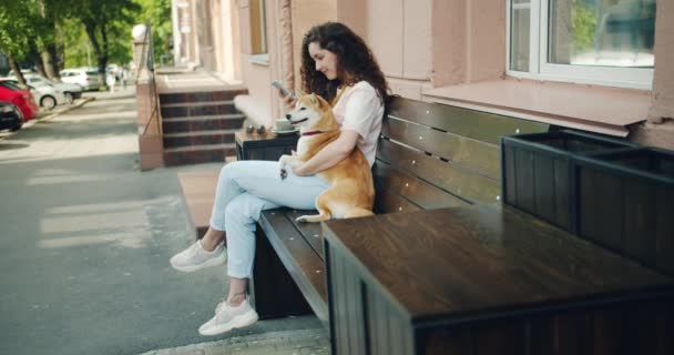 微笑的女孩使用智能手机和拥抱shiba inu狗在咖啡馆户外 — 图库视频影像