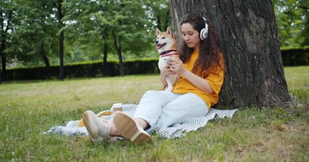 Mulher usando smartphone ouvindo música em fones de ouvido no parque verde com cão — Vídeo de Stock