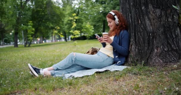 Леди в наушниках наслаждается музыкой, используя смартфон, пьющий кофе в парке — стоковое видео