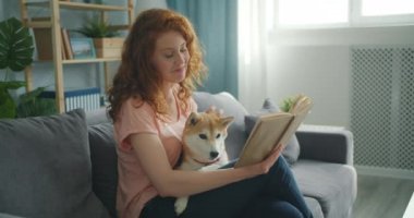 Gülümseyen kız kanepede oturan ve sevimli köpek yavrusu petting evde kitap okuma