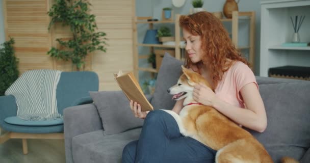 Chica sonriente leyendo historia divertida y acariciando cachorro sentado en el sofá en casa — Vídeo de stock