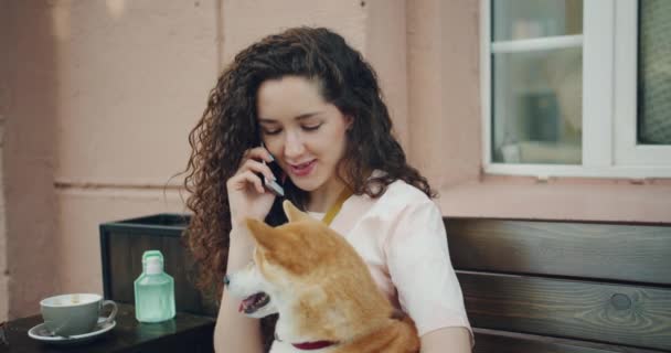 Cámara lenta de la chica hablando en el teléfono móvil y abrazando perro mascota en la cafetería al aire libre — Vídeos de Stock