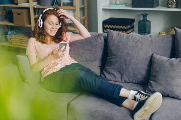Usmívající se dívka naslouchající hudbě přes sluchátka s použitím smartphone doma — Stock fotografie