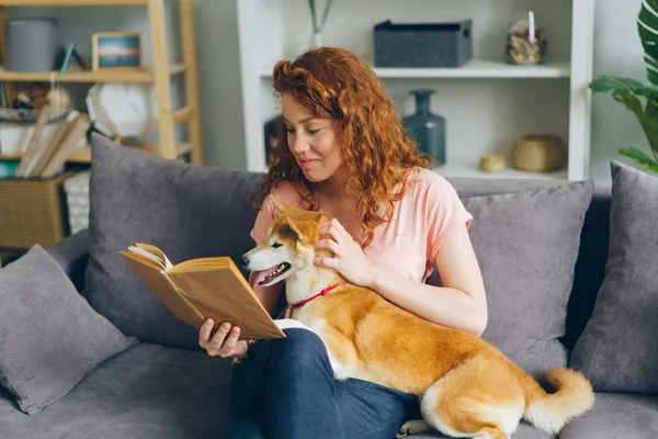 微笑的女孩读有趣的故事和爱抚小狗坐在沙发上在家里 — 图库照片
