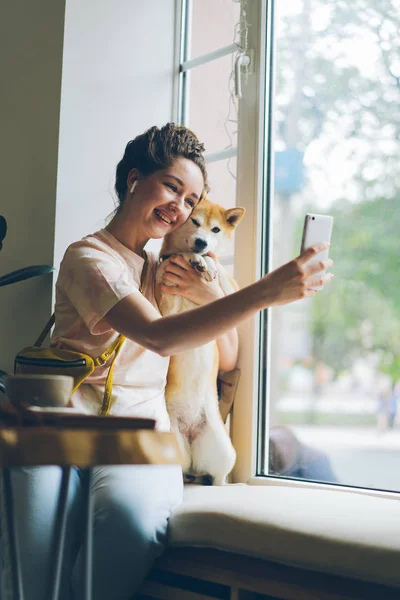 즐거운 소녀 촬영 셀카 에 카페 포옹 강아지 를 사용하여 스마트 폰 에 창틀 — 스톡 사진