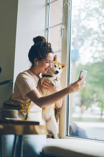 Šťastná dáma s psím, která používá smartphone s kamerou na okenní parapet v kavárně — Stock fotografie
