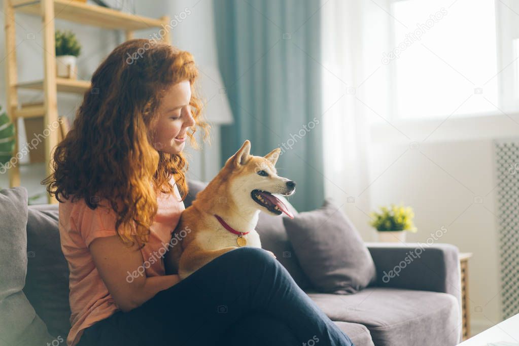 Pretty redhead woman hugging doggy sitting on couch in apartment smiling