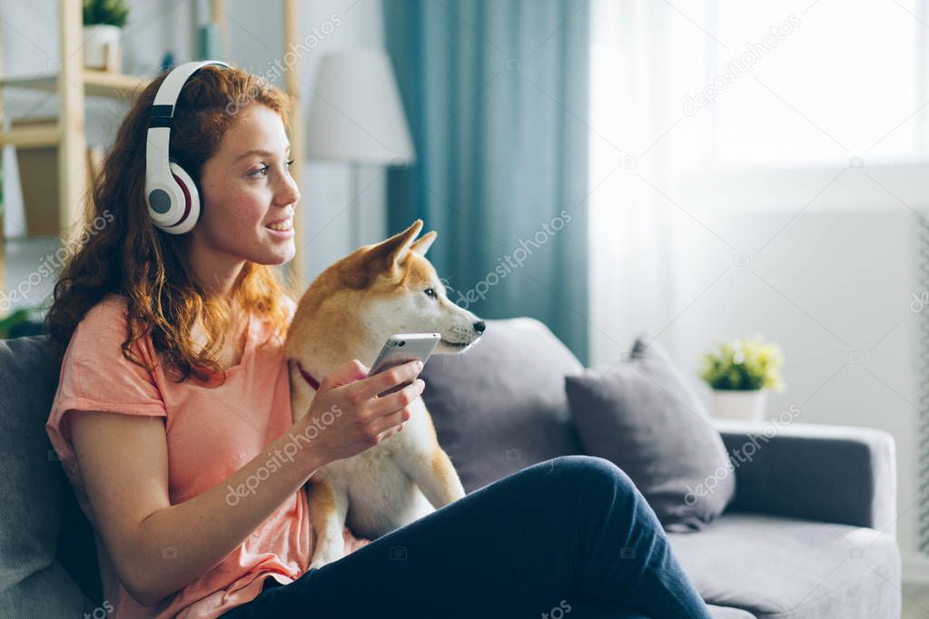 Woman in headphones listening to music petting dog on couch holding smartphone