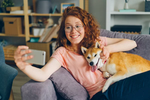 Schönes Mädchen macht Selfie mit Welpen, die zu Hause auf dem Sofa sitzen und das Smartphone benutzen — Stockfoto