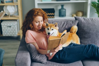 Mutlu genç kadın zevk kitap ve okşayarak sevimli köpek oturma üzerinde kanepe üzerinde düz