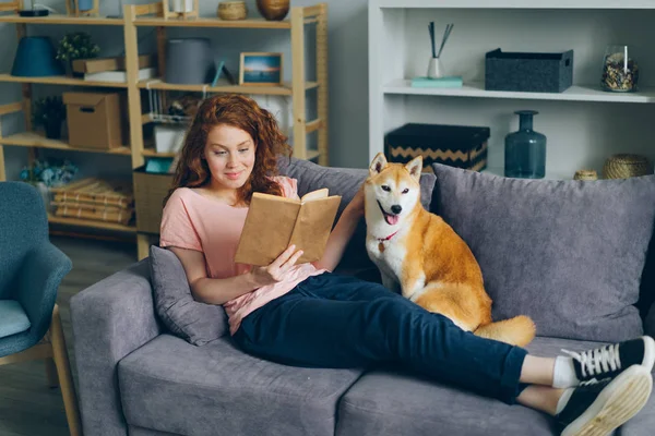 Όμορφη νεαρή γυναίκα διαβάζει βιβλίο και χαϊδεύει το σκυλί κάθεται στον καναπέ στο σπίτι — Φωτογραφία Αρχείου