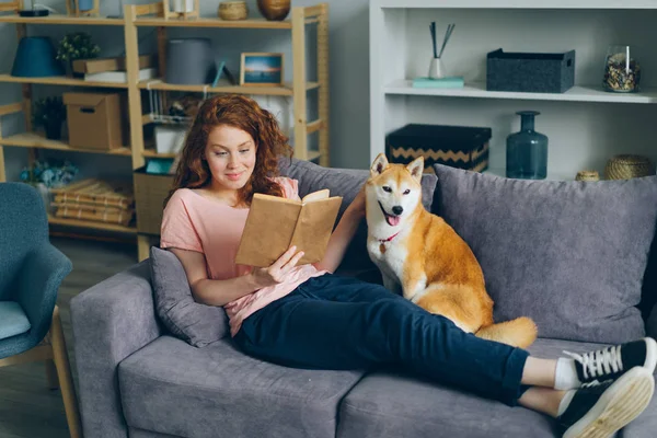 Söt ung kvinna läser bok och smekande hund sitter på soffan hemma — Stockfoto