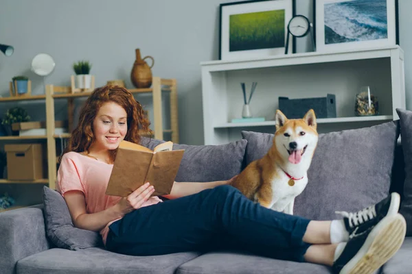 Çekici kız kitap okuma ve düz kanepe üzerinde Shiba inu köpek yavrusu okşayarak — Stok fotoğraf