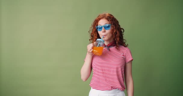 Радісна дівчина в сонцезахисних окулярах п'є апельсиновий сік і посміхається на зеленому фоні — стокове відео