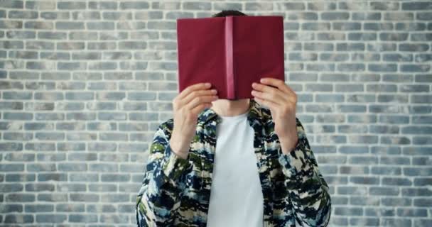 Portrait d'un homme joyeux caché face derrière un livre souriant sur fond de mur de briques — Video