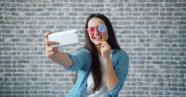 Retrato de una chica bonita tomando selfie con cámara de teléfono inteligente sosteniendo piruleta — Vídeos de Stock