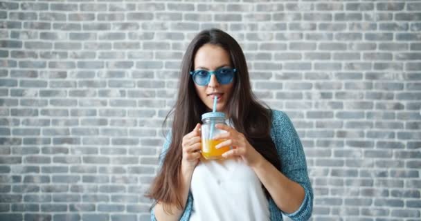 Hübsche Dame mit Sonnenbrille trinkt Orangensaft lächelnd auf Backsteinmauer Hintergrund — Stockvideo