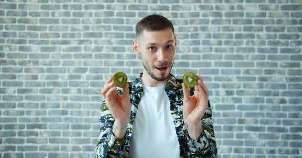 Портрет красивого мужчины с фруктами киви на глазах, улыбающегося на кирпичном фоне — стоковое видео