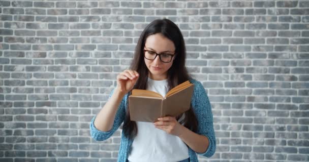 Серйозна дівчина в окулярах читає книгу, що повертає сторінку на фоні цегляної стіни — стокове відео