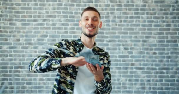 Портрет молодого человека, бросающего деньги и улыбающегося на фоне кирпичной стены — стоковое видео