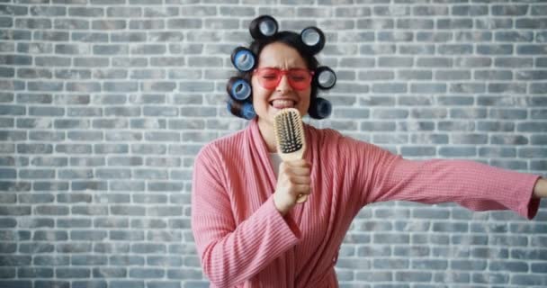 Woman with hair curlers, glasses and bathrobe singing in hairbrush having fun — Stock Video